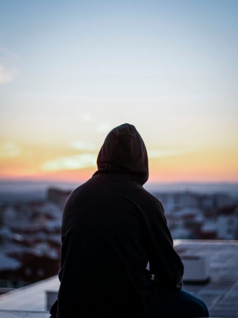 Person mit Kapuze sitzt auf einem Dach und betrachtet einen Sonnenuntergang.